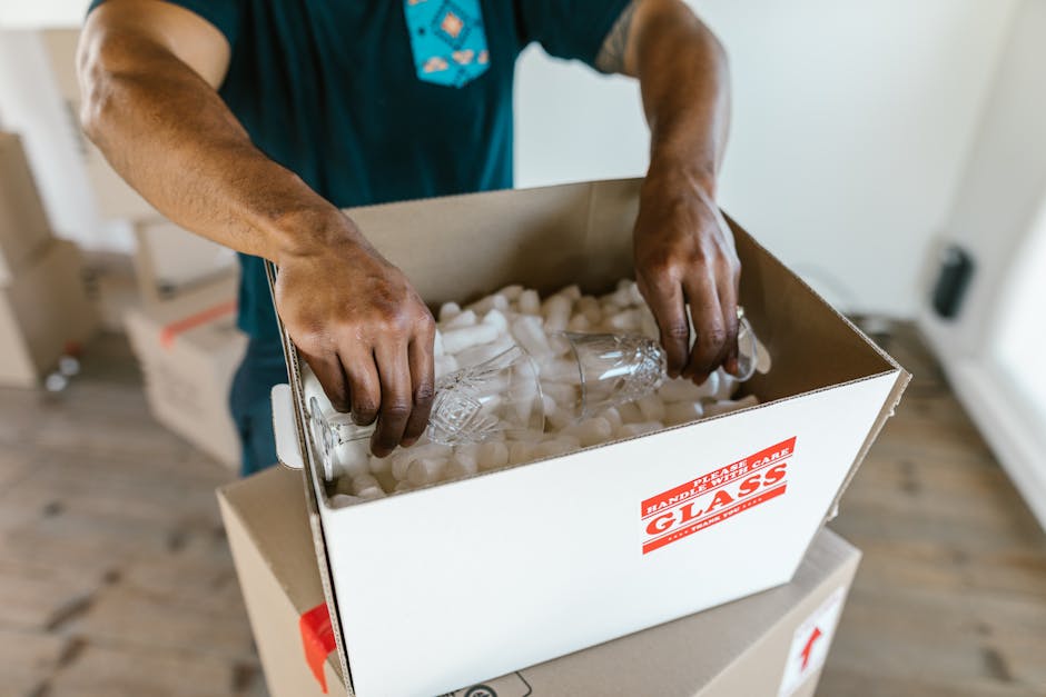 A person wearing a beige long-sleeved top is preparing for a home relocation by packing fragile items. They are placing a white ceramic plate onto a piece of protective bubble wrap on a wooden kitchen